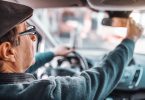 Senior man with hat and eyeglasses adjusting rear mirror while sitting in his car. Other hand on steering wheel. Picture taken from back seat.